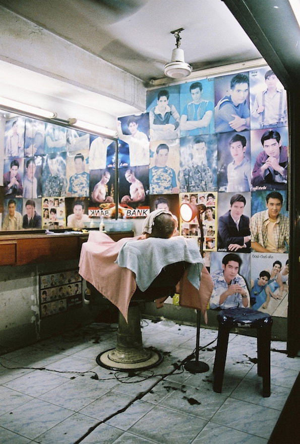 A person sits in a vintage barber chair, draped in a pink and white cape. The walls are covered in a grid of blue-toned portraits of a person in various poses.