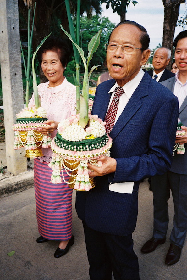 A person in a blue suit and tie holds a traditional Thai floral arrangement. Beside them, another person in a pink lace top and striped skirt holds a similar arrangement.