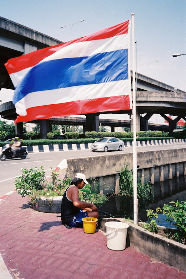 The Thai national flag flies from a tall white pole against a clear blue sky. Below, a person in a black top and white cap squats by a concrete canal edge next to two plastic buckets.