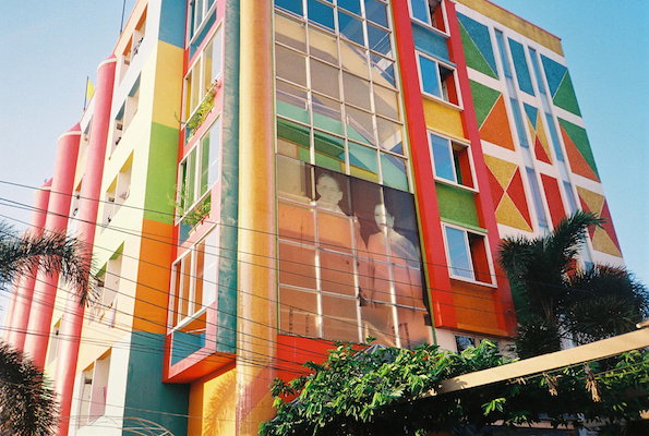 A multi-storey building is decorated in a bold, colourful pattern of geometric blocks and stripes. A large, translucent image of two people is displayed across the central glass facade.