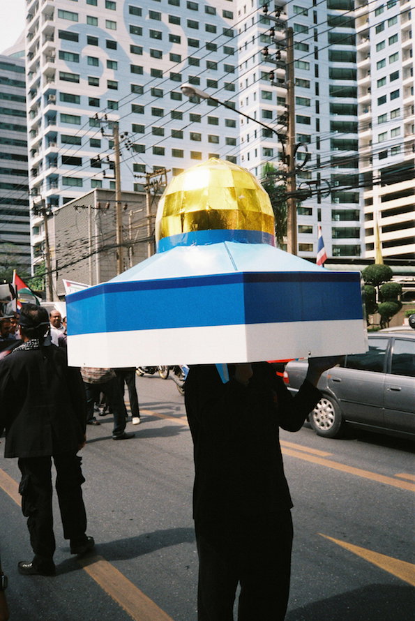 A person stands in a city street wearing a large, geometric headpiece. The structure consists of a wide blue and white base topped with a gold dome.
