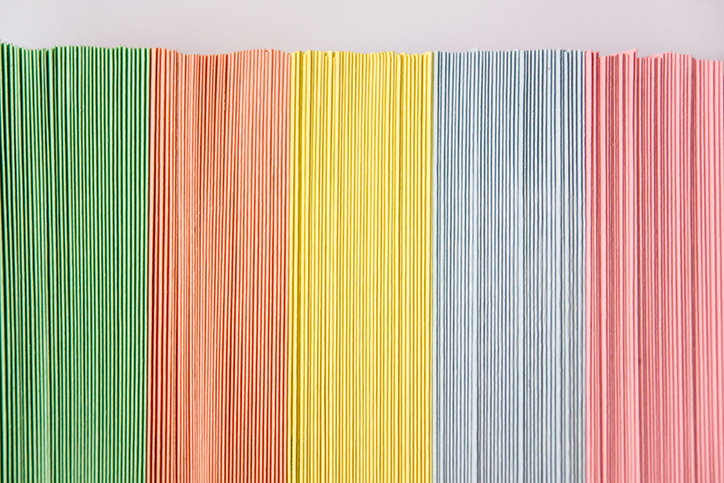 Five vertical bands of corrugated cardboard in green, orange, yellow, blue and pink. The ribbed texture creates a series of fine, parallel lines across each colour.