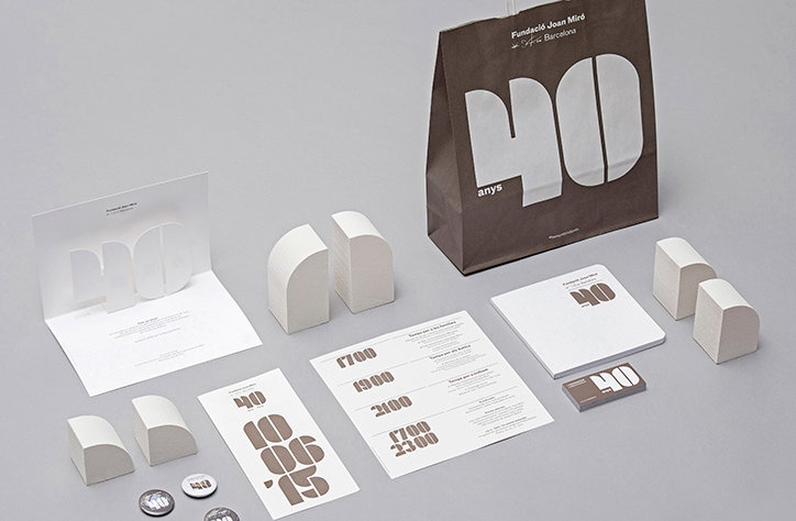A branding suite for the “Fundació Joan Miró” in Barcelona, featuring a brown paper bag with a large white “40 anys”. The set includes a white card, a schedule, and several small, white 3D geometric blocks.