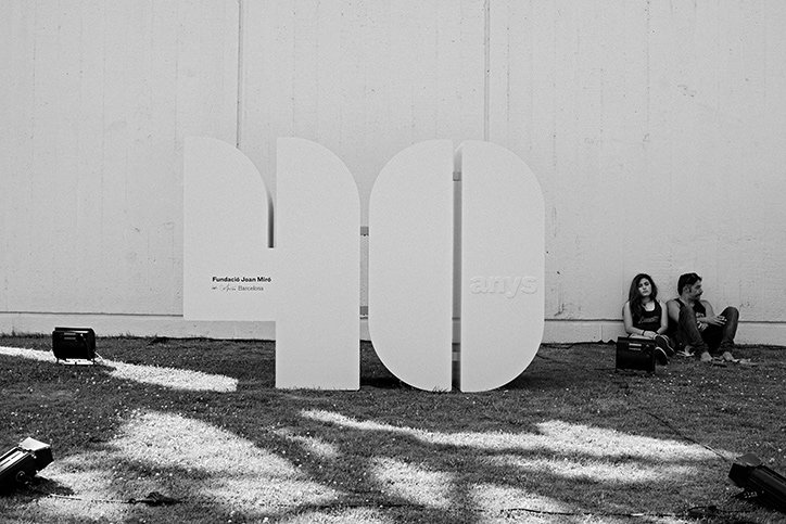 A large, white 3D sculpture of the number “40” stands on a lawn against a plain wall. To the right, two people sit on the grass beside the sculpture.
