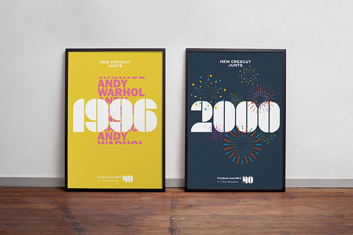 Two framed posters lean against a white wall on a wooden floor. The left poster is yellow with the text “ANDY WARHOL” and “1996”, while the right is dark blue with “2000” surrounded by colourful fireworks. Both feature the heading “HEM CRESCUT JUNTS” and the Fundació Joan Miró logo at the bottom.