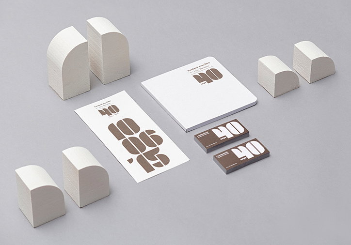 A collection of white branding materials and geometric blocks arranged on a grey surface. A flyer and a square book feature bold typography reading “40 anos”, alongside two small brown boxes with the same text.