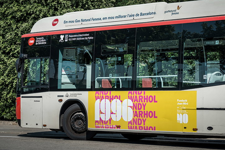 A white city bus features a bright yellow advertisement on its side. The graphic displays the name “Andy Warhol” repeated in pink and orange, with the year “1996” in large white numerals.