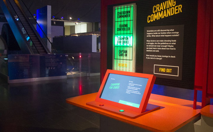 An interactive museum display titled “Craving Commander”. A digital screen on an orange plinth shows the text “Champion for Choice” alongside a list of options.