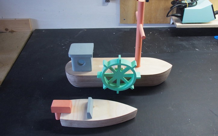 Two wooden toy boats sit on a dark workbench. The larger boat features a teal steering wheel, a light blue block, and a tall orange mast.