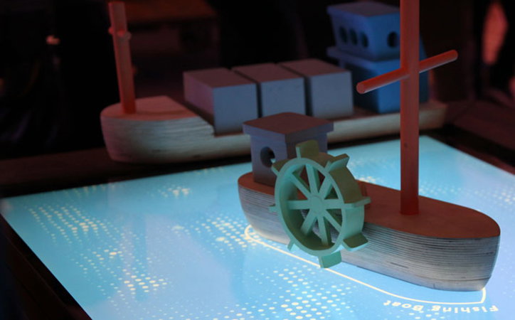 Two wooden toy boats sit on a glowing blue digital surface. The boat in the foreground features a steering wheel and the words “Fishing Boat” printed on the screen beneath it.