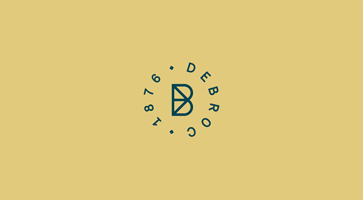 A dark blue logo on a mustard yellow background. A stylised letter “B” is encircled by the word “Debroc” and the numbers “1876”.