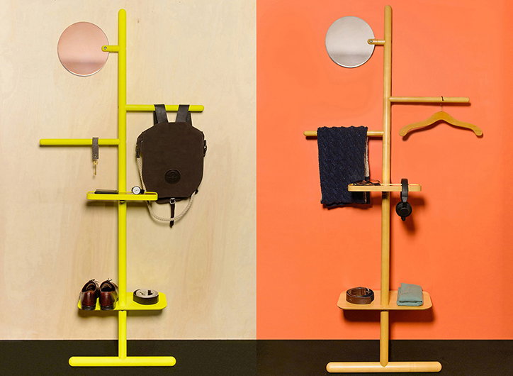 Two minimalist clothing racks stand against contrasting pale wood and orange backgrounds. The yellow rack on the left holds a brown backpack, keys and shoes, while the natural wood rack on the right holds a dark blue garment, headphones and a belt.