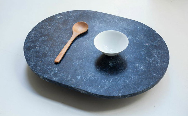 A small white bowl and a wooden spoon rest on a dark, oval marble plinth. The stone surface is mottled with grey and white veins against a deep charcoal background.