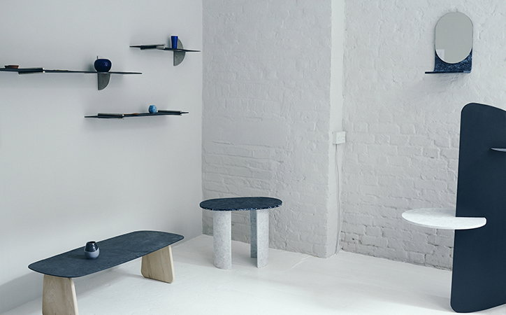 A collection of minimalist furniture and shelving in a bright, white room. The pieces feature dark, organic shapes paired with light-coloured stone and wood supports.