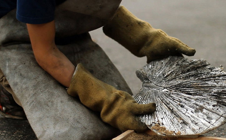 A person wearing heavy yellow work gloves holds a large, charred piece of wood. The wood is silvered and cracked, resembling a burnt shell or a cross-section of a tree.