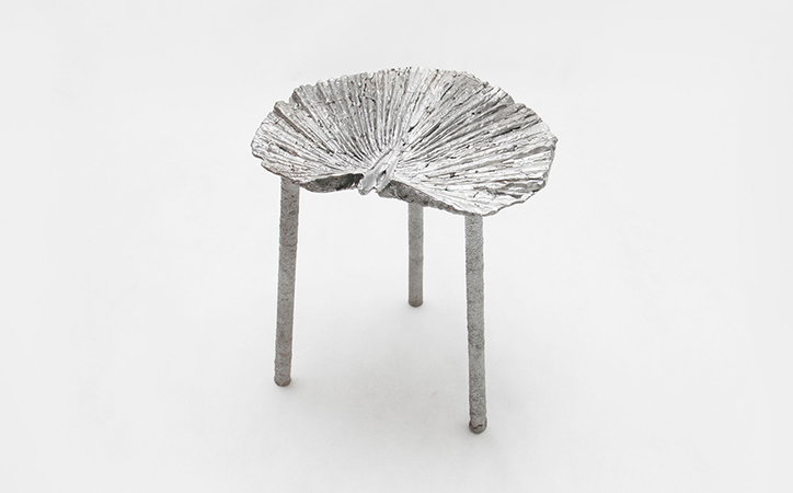 A silver sculpture of a small table with three thin legs. The tabletop is shaped like a textured, ribbed leaf against a plain white background.