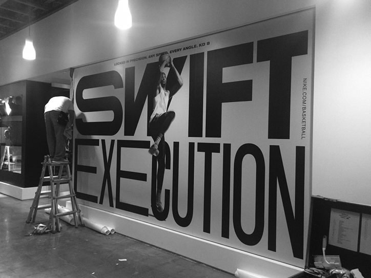 A person on a ladder installs a large black and white Nike advertisement on a white wall. The poster features a basketball player and bold typography that reads “SWIFT EXECUTION”.