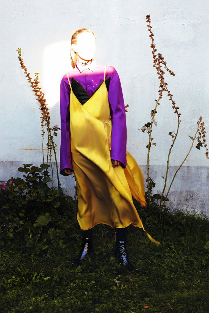 A person stands against a white wall, wearing a purple long-sleeved shirt under a yellow satin slip dress. Their face is obscured by a bright, white glare of light.