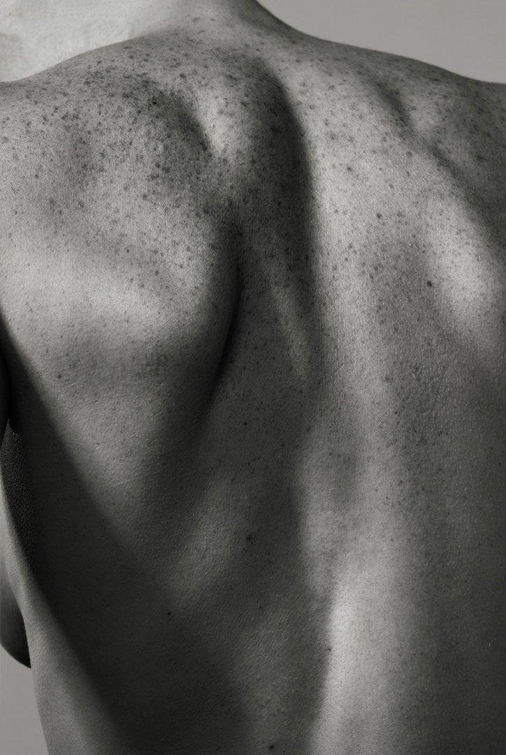 A black and white close-up of a person’s bare back. The light catches the curve of the spine and the texture of freckles across the skin.