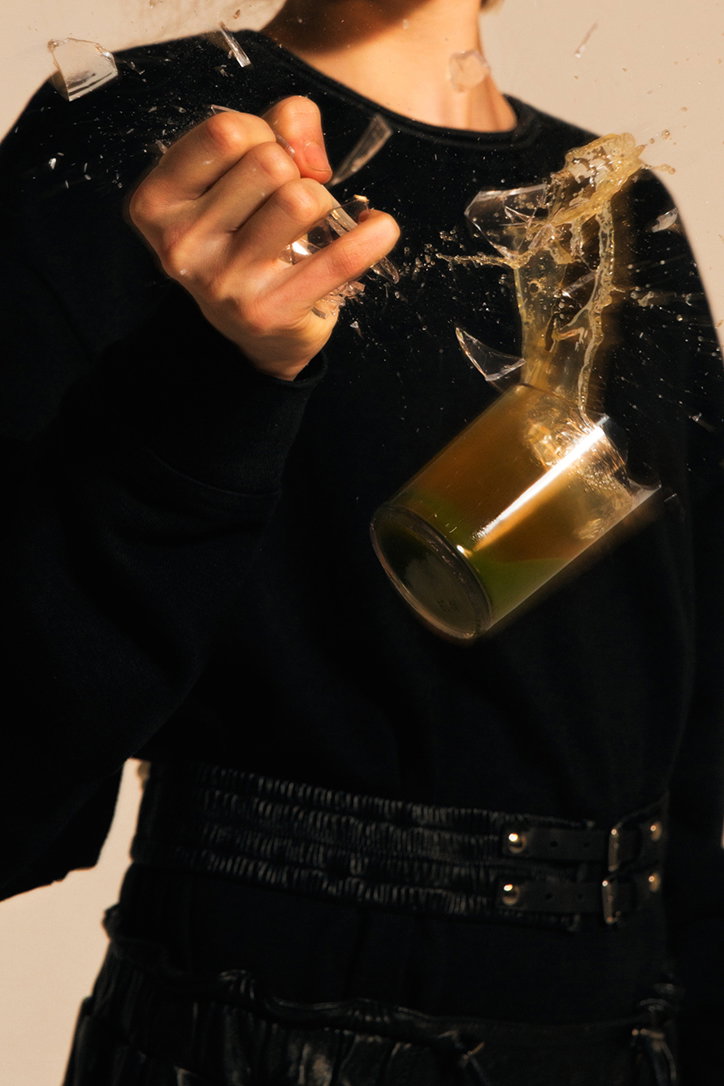 A person in a black sweatshirt holds their hand clenched as a glass shatters in mid-air. Shards of glass and a golden liquid spray across the frame.