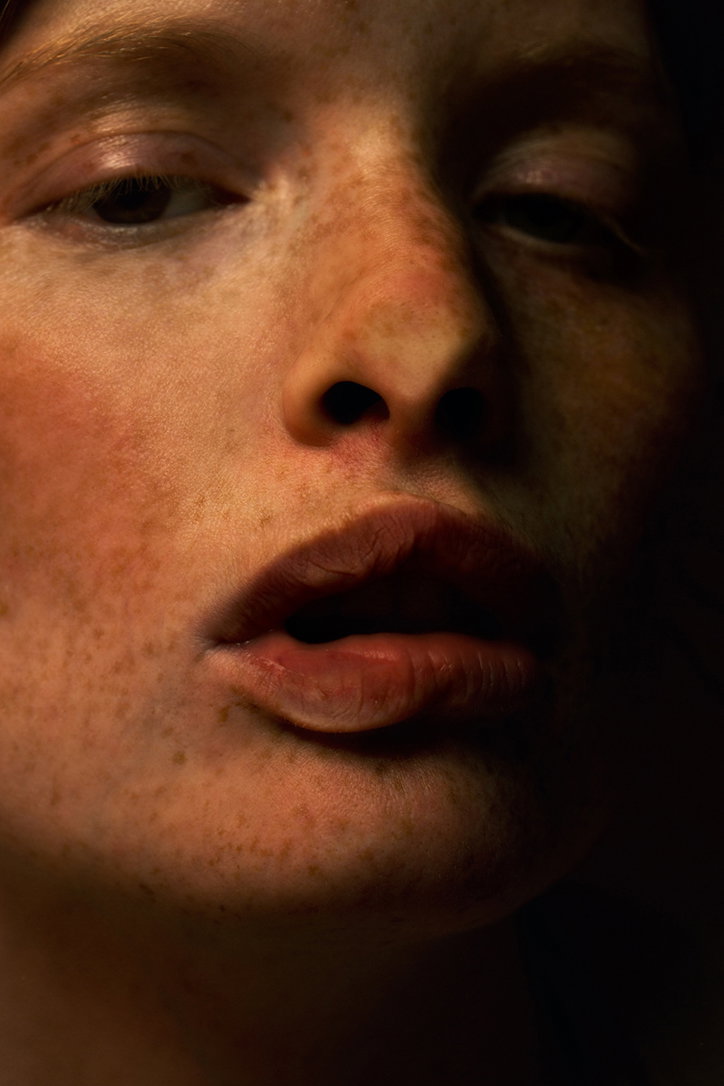 An extreme close-up of a person’s face, focusing on their nose and open mouth. Strong side-lighting casts half the face in deep shadow, highlighting freckles across the skin.