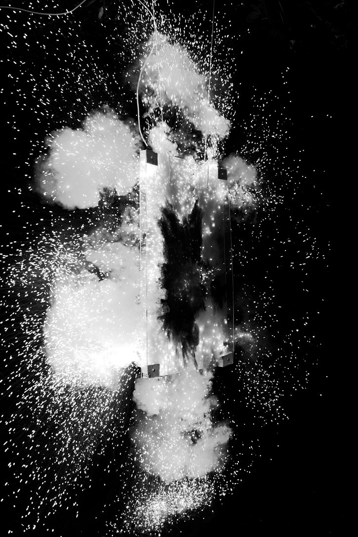 A black and white photograph of a rectangular glass panel shattering. Bright sparks and clouds of white smoke explode outwards against a dark background.