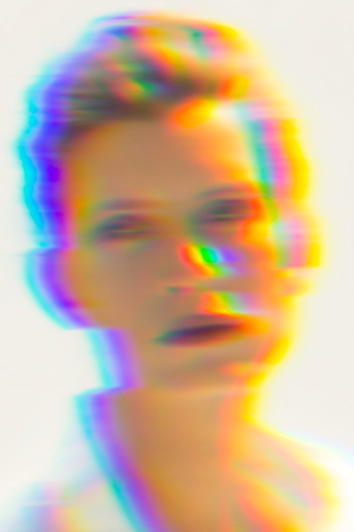 A blurred portrait of a person, distorted by a vibrant chromatic aberration effect. Streaks of neon blue, yellow and red shift across the figure against a plain white background.