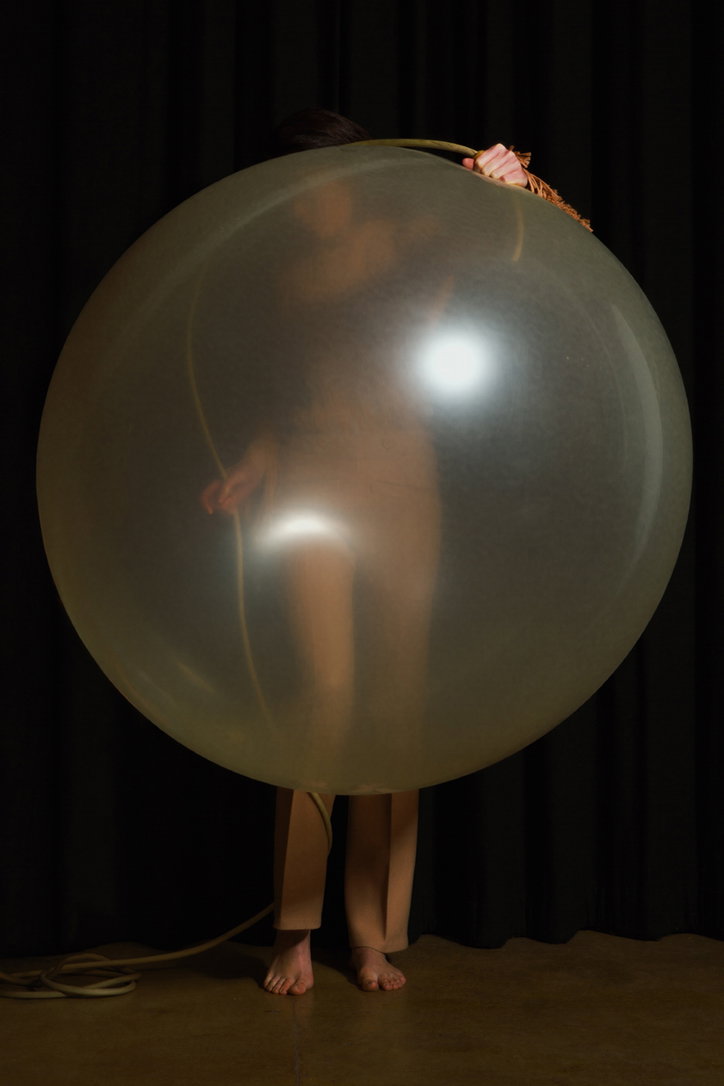 A person stands behind a large, translucent balloon that obscures most of their body. They hold the top of the balloon against a dark curtain backdrop.