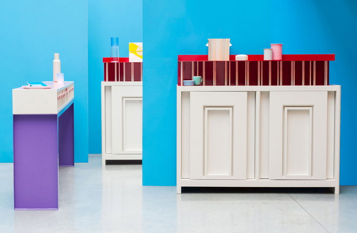 Three minimalist cabinets stand against a bright blue wall. The furniture features a mix of white, purple and red panels, with small ceramics and glassware arranged on top.