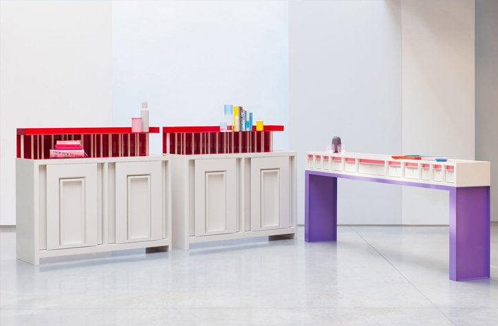 Two white storage units with red shelving stand beside a long, purple-legged table. Various colourful bottles and containers are arranged across the surfaces in a bright, minimalist room.