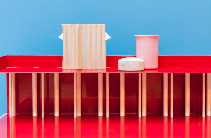 A red shelf holds a closed book, a small marble dish and a pink cup. The objects sit against a bright blue background.