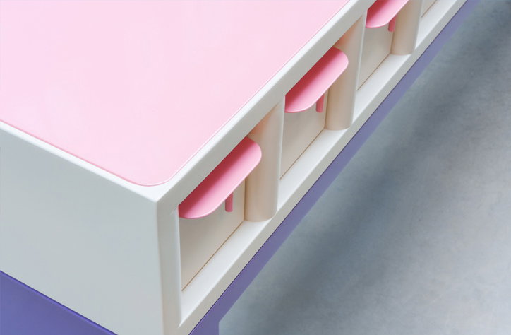 A close-up of a piece of furniture with a pale pink top and a white base. Small, pink semi-circular tabs protrude from a row of recessed slots along the side.