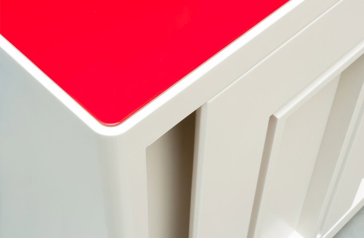 A close-up of a piece of furniture with a glossy red top and a white base. The white surface features a series of deep, vertical recessed grooves.