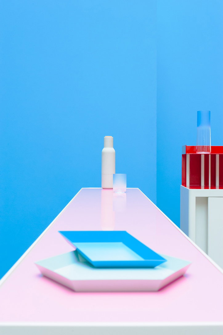 A long pink table holds a blue square tray resting on a larger white hexagonal tray. In the background, a white bottle and a blue glass stand against a bright blue wall.