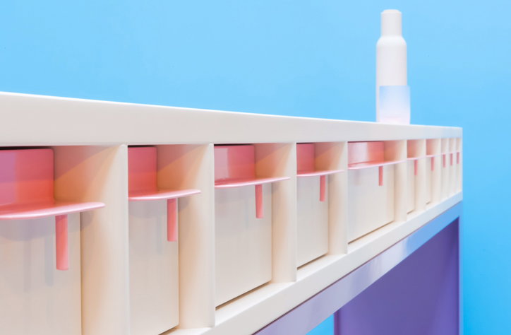A white skincare bottle stands on a long, cream-coloured shelf against a bright blue background. Below the shelf, a row of matching cream compartments each hold a pink product.