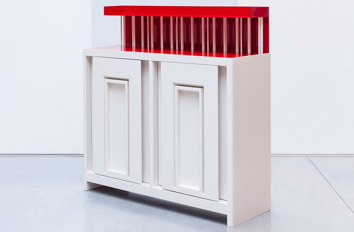 A white wooden sideboard with two paneled doors. A translucent red acrylic structure sits on top, consisting of a flat slab supported by vertical ribs.