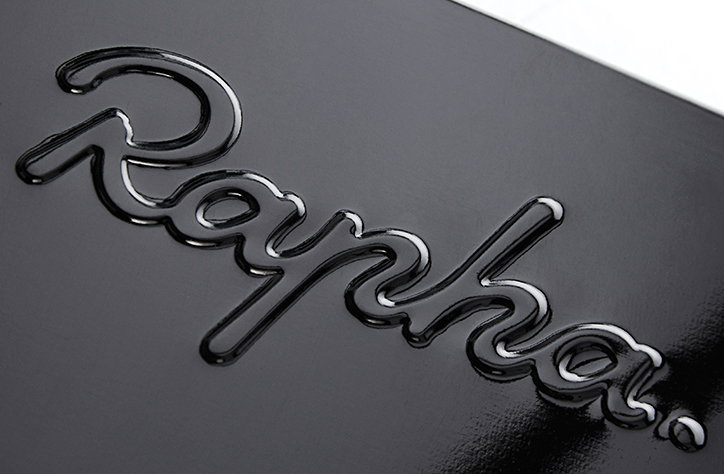 A close-up of the Rapha logo embossed in a glossy black finish. The word “Rapha.” is rendered in a fluid, cursive script that catches the light.