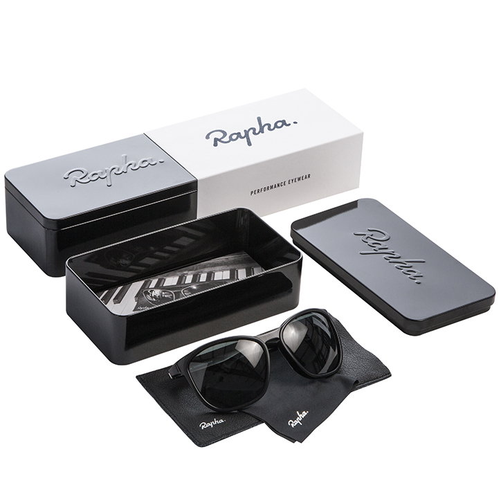 A pair of black sunglasses rests on a black cleaning cloth. Beside them are a black hard case and a matching box, both featuring the “Rapha” logo.
