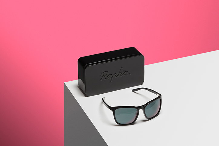 Black sunglasses sit on a white surface next to a black hard case. The case features the embossed “Rapha” logo and is set against a bright pink background.