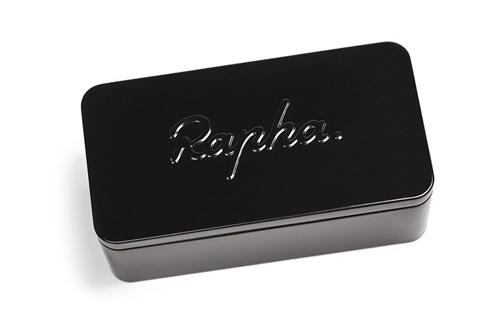 A glossy black rectangular tin set against a plain white background. The word “Rapha” is embossed in a cursive script across the lid.