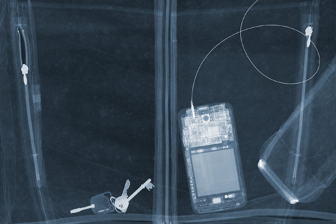An X-ray image of the contents of a bag. Inside, a smartphone with wired earphones, a set of keys and two zips are visible against a dark blue background.