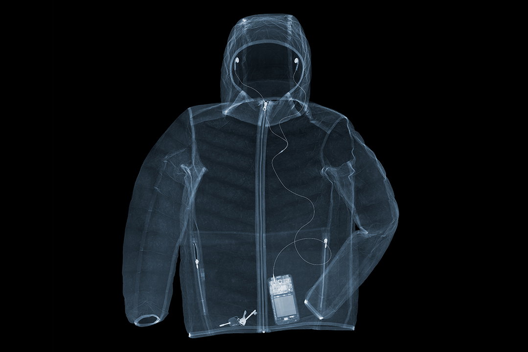 An X-ray of a hooded jacket against a black background. Inside a pocket, a small digital music player is visible, with wires leading up to earbuds in the hood.
