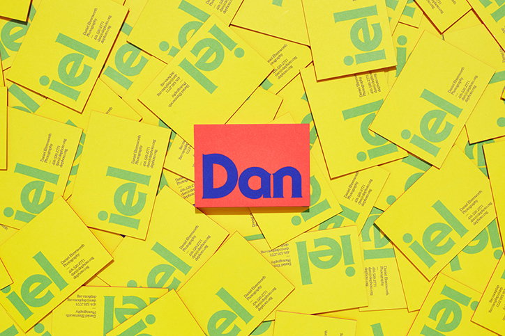 A small red card with the name “Dan” in bold blue type sits atop a pile of yellow business cards. The yellow cards feature a large, green, stylised logo and the name “Daniel Ebenworth”.