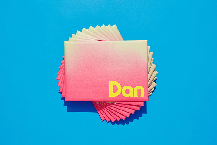 A pink and yellow gradient envelope reads “Dan” in bold yellow type. It sits atop a fan of folded pink and yellow paper against a bright blue background.