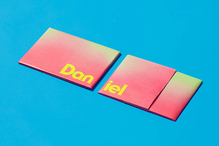 Two gradient booklets in pink and yellow lie on a bright blue surface. The word “Daniel” is printed across them in bold yellow type.