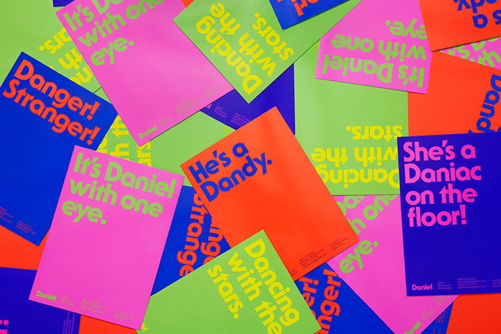 A pile of brightly coloured posters in neon pink, orange, blue and green. The posters feature bold typography with phrases including “It’s Daniel with one eye.”, “He’s a Dandy.” and “She’s a Daniac on the floor!”.