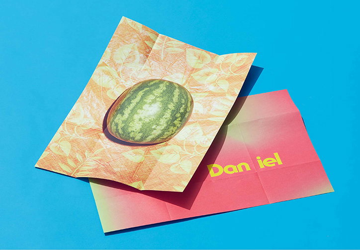 A small watermelon rests on a piece of folded, orange-patterned paper. This sits atop a red gradient envelope that reads “Dan iel” in yellow text, all set against a bright blue background.