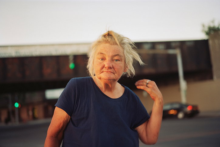 A person with wind-swept blonde hair looks directly at the camera with a neutral expression. They wear a navy blue T-shirt and stand against a blurred urban backdrop of a road and a dark overpass.
