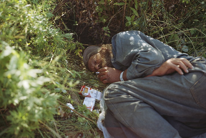 A person lies curled on the ground amidst dense green foliage and undergrowth. They wear a grey garment and a brown head covering, holding a cigarette in their mouth.