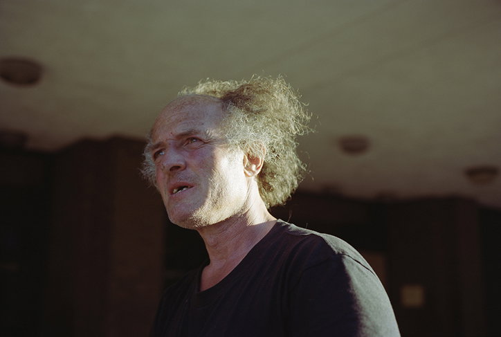 A person with wild, frizzy hair and a receding hairline looks off-camera with a focused expression. They wear a plain black T-shirt and are lit from the side against a muted, architectural background.